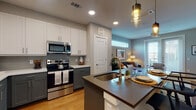 This single-floor residence offers a thoughtfully designed layout that balances modern aesthetics with everyday functionality. The open-concept kitchen is a standout feature, showcasing two-tone cabinetry with white upper cabinets and slate-gray lower cabinets, a glass tile backsplash, white quartz countertops, and a full suite of stainless steel appliances including a French door refrigerator, electric range with microwave, and dishwasher. A large central island with a dark quartz countertop and an undermount sink serves as both a prep space and casual dining area, illuminated by a stylish pendant light. Warm hardwood flooring flows seamlessly throughout the entire residence. The living area is bright and inviting, benefiting from a full-length glass door and a clerestory window that allow abundant natural light to fill the room. A ceiling fan adds comfort overhead. The bedroom is generously proportioned, featuring a ceiling fan, large windows, and easy access to a private en-suite bathroom. The bathroom is well-appointed with a slate-gray vanity, white countertop, a large mirror, and a glass-enclosed walk-in shower. An adjacent walk-in closet provides ample storage. Overall, the space presents a clean, contemporary design with a warm and comfortable ambiance.