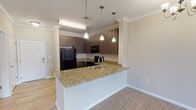 Located at 7107 Brandy Run Dr in Mechanicsville, Virginia, this single-floor residence offers a well-designed, open-concept layout that connects the kitchen and living areas seamlessly. The kitchen is appointed with rich dark wood cabinetry, granite countertops, and a full suite of black appliances including a refrigerator, range, microwave, and dishwasher. A peninsula with a built-in sink and pendant lighting above serves as both a functional workspace and a natural divider between the kitchen and the adjacent living and dining areas. Crown molding runs throughout the main living spaces, adding a polished architectural detail. The living area features plush carpeting, a ceiling fan with chandelier-style lighting, and access to a door with sidelights that brings in natural light. Bedrooms are carpeted and generously sized, each equipped with ceiling fans and ample closet space, including a walk-in closet in the primary bedroom. The bathroom features granite vanity countertops, a full tub-shower combination with tile surround, and wood-look flooring. A stacked washer and dryer unit is conveniently located within the bathroom area, providing in-unit laundry. The overall aesthetic is warm and neutral, with consistent finishes throughout that create a cohesive and functional living environment.