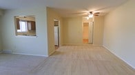 This single-floor residence offers a clean, functional layout designed for comfortable everyday living. The main living area is spacious and open, featuring light beige carpeting that adds warmth underfoot and neutral-toned walls that create a bright, welcoming atmosphere. A ceiling fan with an integrated light fixture is centrally positioned, providing both air circulation and ambient lighting throughout the space.

The kitchen is accessible directly from the living area via an open pass-through cutout in the dividing wall, allowing for a connected, semi-open feel between the two spaces. The kitchen features wooden cabinetry and is equipped with a white refrigerator, contributing to a classic, practical design.

A hallway visible from the living room leads to additional areas of the unit, with what appears to be a tiled entry or bathroom space visible at the end. The transition from carpet in the living area to hard flooring in the connecting spaces adds visual contrast and practical zoning throughout the floor plan.

Overall, the space maintains a straightforward residential character, with consistent neutral finishes, well-proportioned rooms, and an efficient single-level layout that prioritizes ease of movement and livability.