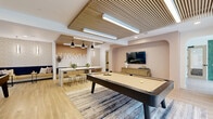 Located at 6220 Guilford Ave in Indianapolis, Indiana, this thoughtfully designed amenity space delivers a refined social environment across a single level. The open layout centers around a main game and lounge area anchored by a full-size billiards table and a sleek foosball table, both set atop a large area rug over light wood-look tile flooring that runs throughout. A striking slatted wood ceiling treatment adds warmth and architectural character, complemented by modern linear pendant lighting over the kitchen bar area and recessed lighting throughout.

The kitchenette features warm natural wood cabinetry, a white marble-look quartz countertop, a stainless steel undermount sink with a brushed gold faucet, a beverage refrigerator, and an eye-catching backsplash of mixed white and tan rectangular tiles. A counter-height bar with stools makes it ideal for casual entertaining.

A dedicated lounge nook with a wall-mounted flat-screen TV is framed by a softly arched accent wall in warm blush tones, paired with a natural wood media console. Floor-to-ceiling sliding glass doors on two sides of the space flood the interior with natural light and provide direct views of and access to the outdoor pool and deck area, seamlessly connecting indoor and outdoor living.