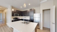 This single-story home offers a thoughtfully organized layout across three bedrooms and two bathrooms. The living area is bright and open, finished with light-toned hardwood-style flooring that extends throughout the main areas. Multiple windows and a glass exterior door flood the space with natural light and provide access to an outdoor balcony, enhancing the airy feel of the interior.

The kitchen is a functional centerpiece, featuring dark espresso-toned cabinetry with ample upper and lower storage, white countertops, a central island with a built-in sink, and stainless steel appliances including a gas range, microwave, and refrigerator. Pendant lighting accents the island, adding a polished touch.

The bedrooms are carpeted for comfort. The primary bedroom is generously sized and includes a walk-in closet outfitted with wire shelving and hanging rods. Secondary bedrooms feature sliding-door closets with integrated shelving.

Both bathrooms are finished with dark cabinetry, white countertops, and light wood-style flooring. One bathroom conveniently incorporates an in-unit washer and dryer with overhead shelving. A dedicated hallway connects all rooms, ensuring a practical and cohesive flow throughout the home.