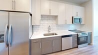 Located at 1803 S 46th St in Rogers, Arkansas, this thoughtfully designed one-bedroom, one-bathroom unit offers a clean, modern aesthetic across its single-floor layout. The galley-style kitchen is a standout feature, equipped with stainless steel appliances including a refrigerator, range, microwave, and dishwasher. White upper cabinetry pairs with gray lower cabinets, complemented by a white subway tile backsplash, white quartz countertops, and warm hardwood-style flooring, creating a contemporary two-tone design. The bedroom is finished with soft carpeting, a ceiling fan, and a window that allows natural light and exterior views to fill the room. The bathroom features hardwood-style flooring, a white subway tile walk-in shower with brushed nickel fixtures, a single-sink vanity with white countertop, and a mirror. A dedicated laundry area houses a stacked front-loading washer and dryer unit, adding everyday convenience. Throughout the space, neutral white walls, recessed lighting, and consistent flooring choices create a cohesive, bright, and functional living environment well suited for comfortable single-occupant living.