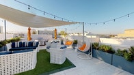 This vibrant rooftop terrace is a thoughtfully designed outdoor retreat that combines modern aesthetics with comfortable, functional amenities. The open-air deck is finished with light-toned terrazzo-style tile flooring and features a central artificial turf seating area that anchors the main lounge zone. White lattice-style outdoor furniture with navy blue cushions is arranged to encourage social gathering, while a pair of white wicker egg swing chairs with bold orange cushions serve as standout accent pieces throughout the space. A large sail shade canopy stretches overhead, providing relief from direct sunlight, and strands of bistro string lights crisscross above, creating an inviting ambiance for evening use. Orange market umbrellas add pops of color near the BBQ grill station, which is equipped for outdoor cooking and entertaining. Raised planter beds filled with drought-tolerant succulents and agave line the perimeter, lending a clean, contemporary landscape element. The terrace is accessible via elevator and a gated entry passage. Panoramic views of the surrounding cityscape, palm trees, and distant mountain ranges are visible from every angle, making this rooftop an exceptional space for both daytime relaxation and evening social events.