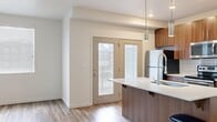 This well-designed single-floor unit offers a thoughtful layout across a comfortable footprint. The open-concept kitchen and living area serves as the heart of the space, where the kitchen stands out with warm walnut-toned cabinetry, a white quartz waterfall island with an undermount sink and professional-style faucet, white subway tile backsplash, and a full suite of stainless steel appliances including a refrigerator, electric range, and microwave. Pendant lighting above the island adds a modern touch, while wood-style plank flooring and a glass-paneled exterior door bring in natural light and outdoor views. The living area benefits from a ceiling fan and the same durable flooring, lending continuity to the open layout. The bedroom is a calm, carpeted retreat with a window providing natural light and a dedicated walk-in closet featuring built-in hanging rods and shelving for ample storage. The bathroom mirrors the kitchen's design aesthetic, showcasing a walnut vanity with white quartz countertop, a large mirror, and wood-style flooring, creating a clean, cohesive finish throughout the unit.