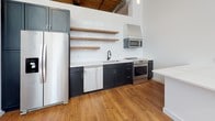 Located at 220 Merrimack in Lawrence, MA, this loft-style unit beautifully marries industrial character with contemporary design. The open-plan living area is anchored by striking exposed brick walls and soaring wood-beam ceilings with visible ductwork, lending an authentic warehouse aesthetic. Warm hardwood floors run throughout, reflecting abundant natural light that pours in through large, multi-pane factory-style windows, offering views of the surrounding area.

The kitchen is well-appointed with dark navy cabinetry, crisp white quartz countertops, and a classic white subway tile backsplash. Open wood shelving adds both function and warmth, while stainless steel appliances — including a range with hood, dishwasher, microwave, and refrigerator — provide a fully equipped cooking space. A central island offers additional counter space and storage.

The bathroom continues the refined material palette with hexagonal tile flooring featuring decorative mosaic accents, subway tile walls, a vessel sink atop a wood vanity with gold fixtures, and a tall storage cabinet that maximizes functionality. The overall ambiance of the unit is one of spacious, light-filled industrial sophistication — honoring the building's historic mill origins while delivering a polished, modern living experience.