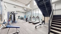 This fitness center is thoughtfully designed across two distinct zones, offering a comprehensive workout experience. The main exercise area is an open, industrial-inspired space characterized by soaring ceilings, expansive floor-to-ceiling windows, and clerestory windows that flood the room with abundant natural light and views of the surrounding exterior. The polished concrete-style flooring, exposed structural columns, and overhead ceiling fans contribute to the airy, loft-like atmosphere. This primary zone is well-appointed with a full lineup of cardio machines — including treadmills, ellipticals, and a rowing machine — alongside a dedicated free weights section featuring a complete dumbbell rack, weight bench, punching bag, stability balls, and a large wall mirror. A bold athletic mural and staircase accent add visual interest to the space. The secondary zone offers a more intimate studio setting, finished with warm wood-style plank flooring and rich wood-paneled walls with frosted glass insets. This room houses cycling bikes, kettlebells, a dumbbell rack, and a large wall-mounted display screen, making it ideal for group or individual training sessions. Together, the two areas provide a versatile and well-rounded fitness facility.