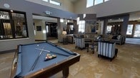 This elegantly designed amenity lounge offers a sophisticated and inviting atmosphere perfect for recreation and relaxation. The space is anchored by a full-size billiards table with slate-blue felt, positioned prominently on warm travertine tile flooring that extends throughout the room. Soaring ceilings with clerestory windows flood the interior with natural light, creating an airy, open ambiance that feels expansive and welcoming.

The lounge area is thoughtfully furnished with a large sectional sofa, striped accent chairs, and sleek dark-framed coffee and side tables, all arranged to encourage conversation and comfort. Decorative artwork, including a bold abstract blue canvas, adds a curated, contemporary touch to the neutral color palette of grays, taupes, and creams.

A dedicated desk or gaming station is visible in the background, offering additional functionality. The space also features a separate adjacent room visible through a wide opening, hinting at further amenities or activity areas beyond. Architectural details such as recessed lighting, decorative columns, and layered ceiling heights contribute to the refined, upscale character of this communal space, making it an ideal gathering spot for leisure and social interaction.