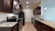 This single-floor residence offers a thoughtfully designed layout that balances functionality and style throughout its living spaces. The galley-style kitchen serves as a central highlight, featuring rich dark espresso cabinetry paired with light granite countertops that provide a striking contrast. Stainless steel appliances, including a range, microwave, refrigerator, and dishwasher, equip the kitchen for everyday use. Light wood-look flooring runs the length of the kitchen, complementing the overall aesthetic. A pass-through or peninsula connects the kitchen to the open living area, which is illuminated by pendant lighting and recessed fixtures. The living space is generously proportioned, featuring the same wood-look flooring and a ceiling fan overhead. French doors with a transom window open to an outdoor area, allowing natural light to flow into the room. Bedrooms are carpeted for comfort and include ceiling fans and windows that bring in additional natural light. A dedicated in-unit laundry area with a full-size washer and dryer is conveniently accessible directly from the kitchen, adding practical value to the home's overall design.