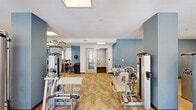 Located at 1223 N Haven Dr in Fayetteville, Arkansas, this fitness center offers a generously sized, single-floor exercise space designed to support a comprehensive workout experience. The room is thoughtfully laid out to accommodate a wide variety of training modalities without feeling cramped.

The flooring throughout features a distinctive light wood herringbone pattern, lending the space a clean and polished aesthetic. The white ceiling is punctuated by multiple recessed skylight panels that flood the room with bright, even illumination, complemented by large windows along one wall that bring in natural light and views of the surrounding greenery.

The space is outfitted with professional-grade equipment, including multiple Cybex strength machines covering a range of muscle groups, True treadmills, and an elliptical trainer. A dedicated functional training area includes resistance cables, stability balls, resistance bands, and wall-mounted medicine balls, catering to flexibility and dynamic workout routines. A dumbbell rack is also visible along one wall, completing a well-rounded selection of free-weight options.

Accent walls in a muted steel blue add a calm, focused ambiance to the space, while structural columns are finished in a matching tone, reinforcing the cohesive color palette. The layout provides clear circulation paths between equipment zones, making the facility practical and user-friendly for daily exercise routines.
