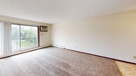 Located at 855 County Road E in Vadnais Heights, Minnesota, this single-floor unit offers a practical and comfortable layout designed for everyday living. The spacious living area is finished with plush wall-to-wall carpeting, neutral-toned walls, and a ceiling fan with integrated lighting, creating a bright and airy atmosphere. A wall-mounted air conditioning unit and baseboard heating provide year-round climate control. A sliding glass door opens to a private balcony, offering pleasant outdoor views of the surrounding greenery. The living space transitions seamlessly into the kitchen, which features warm wood cabinetry extending to the ceiling for generous storage, a tile-style floor, a coil-top electric range, a refrigerator, and a sink with ample counter space. A walk-in closet is conveniently accessible directly from the kitchen area. The bedroom is well-proportioned, featuring carpeted flooring, a ceiling fan, a window with natural light, and baseboard heating. The full bathroom is finished with classic white tile in the tub and shower surround, a tiled floor, a wooden vanity with under-sink storage, and a toilet. Throughout the unit, dark wood trim and doors provide a cohesive, traditional aesthetic, while textured ceilings add a classic touch to each room.