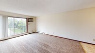 Located at 855 County Road E in Vadnais Heights, Minnesota, this single-floor unit offers a practical and comfortable layout designed for everyday living. The spacious living area is finished with plush wall-to-wall carpeting, neutral-toned walls, and a ceiling fan with integrated lighting, creating a bright and airy atmosphere. A wall-mounted air conditioning unit and baseboard heating provide year-round climate control. A sliding glass door opens to a private balcony, offering pleasant outdoor views of the surrounding greenery. The living space transitions seamlessly into the kitchen, which features warm wood cabinetry extending to the ceiling for generous storage, a tile-style floor, a coil-top electric range, a refrigerator, and a sink with ample counter space. A walk-in closet is conveniently accessible directly from the kitchen area. The bedroom is well-proportioned, featuring carpeted flooring, a ceiling fan, a window with natural light, and baseboard heating. The full bathroom is finished with classic white tile in the tub and shower surround, a tiled floor, a wooden vanity with under-sink storage, and a toilet. Throughout the unit, dark wood trim and doors provide a cohesive, traditional aesthetic, while textured ceilings add a classic touch to each room.