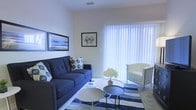 This single-floor space showcases a thoughtfully designed living area that balances comfort with a clean, coastal aesthetic. The living room serves as the focal point, featuring a large sliding glass door fitted with vertical blinds that allow abundant natural light to fill the room while offering privacy control. The neutral, light-toned walls provide a versatile backdrop that enhances the sense of openness throughout the space.

The living area is anchored by a deep navy tufted sofa accented with patterned throw pillows, complemented by a sleek white accent chair that adds a modern contrast. A blue-and-white striped area rug grounds the seating arrangement, reinforcing the room's coastal theme. Two circular white coffee tables paired with a smaller dark side table create a functional and stylish centerpiece for the space.

Wall art featuring ocean-inspired imagery further ties the nautical theme together, while a vase of white florals on the coffee table adds a fresh, decorative touch. The ceiling features a recessed vent, suggesting a central air system for year-round comfort. Overall, the space presents a bright, well-coordinated environment with a relaxed yet polished ambiance.