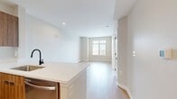 The Delancey at Shirlington Village A1C floor plan offers 540 square feet of efficiently designed living space across one bedroom and one bathroom. Light hardwood flooring flows consistently throughout the entire unit, creating a cohesive and bright aesthetic. The open-concept kitchen and living area serves as the heart of the apartment, featuring warm wood cabinetry, stainless steel appliances including a refrigerator, range, and microwave, a white tile backsplash, and crisp quartz countertops with a breakfast bar peninsula. A matte black faucet and hardware add a contemporary contrast to the neutral palette. The living area is spacious and open, benefiting from a clean, minimalist design with neutral greige walls and ample wall space. The 130 sq ft bedroom is well-proportioned, featuring a large window that brings in natural light and views of the surrounding neighborhood. The bathroom is finished with white subway tile surrounding a soaking tub and shower combination, complemented by matte black fixtures for a modern, polished look. A connecting hallway links the rooms efficiently, completing this thoughtfully designed apartment.