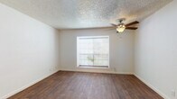 This single-story space is designed with a clean, functional layout that flows through several distinct areas. The main living area features warm wood-look flooring underfoot and a textured popcorn ceiling overhead, complemented by a ceiling fan with a light fixture. A window fitted with horizontal blinds allows natural light to filter in while offering a view of greenery outside.

The kitchen is efficiently arranged in an L-shaped configuration, outfitted with white cabinetry, light-toned countertops, and white appliances including a coil-top electric range, a refrigerator, and a double-basin stainless steel sink. An overhead range hood and under-cabinet lighting add practicality to the workspace.

A hallway connects the living area to the private quarters, with a pass-through window offering a view into the kitchen. The bedroom mirrors the living area in style, featuring the same wood-look flooring, a ceiling fan, a textured ceiling, and a window that brings in natural light.

The bathroom is finished in a neutral palette, featuring a vanity with a speckled countertop, a tiled tub-and-shower combination, and wood-look flooring that maintains consistency with the rest of the space. The overall ambiance is practical and understated, with a neutral color scheme throughout.