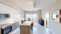 This thoughtfully designed one-bedroom, one-bathroom residence offers a compelling blend of modern aesthetics and functional living. The open-concept layout seamlessly connects the kitchen and living area, anchored by light wood-tone flooring that runs throughout the main space. The kitchen is a standout feature, outfitted with two-tone cabinetry combining walnut-finish lower cabinets and gloss white upper cabinets, quartz countertops, a tiled backsplash, and stainless steel appliances including a built-in microwave, range, and full-size refrigerator. A central island with quartz countertop provides additional prep space and casual seating. The living area is bathed in natural light from expansive floor-to-ceiling windows that frame dramatic, elevated city skyline views. The bedroom continues this visual connection to the city, featuring the same large-format windows that flood the carpeted space with light and panoramic urban scenery. A dedicated closet provides practical storage. The bathroom offers a clean, spa-inspired design with a glass-enclosed walk-in shower clad in grey subway tile, a floating wood-tone vanity, white tile flooring, and built-in open shelving for linens. The overall ambiance is sleek, airy, and sophisticated throughout.