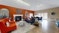 This expansive 990 square foot communal area showcases a harmonious blend of modern design and comfort. The space is characterized by its vaulted ceilings, which create an open and airy atmosphere, enhanced by track lighting that provides both ambient and focused illumination. Large windows and French doors flood the area with natural light, offering views of the surrounding landscape.

The room's color scheme is a striking combination of neutral tones and bold orange accents, which add warmth and energy to the space. A focal point of the room is the white-mantled fireplace, flanked by arched mirrors that reflect light and expand the visual space. The hardwood flooring throughout adds a touch of elegance and continuity.

The furniture arrangement is designed for versatility, featuring a mix of seating options including a plush blue sofa, vibrant orange armchairs, and additional seating areas for dining or work. A glass coffee table and various side tables provide functional surfaces while maintaining a sense of openness. Art pieces, including a large abstract painting and a metallic wall sculpture, add visual interest and sophistication to the decor.

The space is multi-functional, suitable for relaxation, entertainment, and potentially work or study. A small kitchenette or bar area is visible, suggesting the room's capacity for hosting social gatherings. The overall design creates an inviting and dynamic environment that balances style with practicality in this single-floor property.