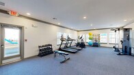 This single-floor amenity facility at 1880 Pat Thomas Pkwy in Quincy, Florida offers a thoughtfully designed space combining fitness and productivity areas. The layout is divided into distinct functional zones, providing residents with a comprehensive community amenity experience.

The fitness area forms the heart of the space, featuring durable carpeted flooring and recessed lighting throughout. It is well-equipped with multiple treadmills, an elliptical cross-trainer, a weight bench, a tiered dumbbell rack with a full range of free weights, and a storage rack holding an assortment of medicine balls and exercise balls in varying sizes. Windows along multiple walls allow natural light to fill the room, creating a bright and energizing workout environment.

An adjoining open area provides additional floor space for stretching or functional training, and a wall-mounted flat-screen television adds to the amenities. This zone connects seamlessly to the rest of the facility through interior doorways.

The business center area is finished with light hardwood-style flooring and decorated with colorful framed artwork, lending it a welcoming and modern aesthetic. A built-in counter-height workstation accommodates multiple users simultaneously, equipped with desktop computers and seating for several chairs. Large windows with blinds offer abundant natural light and views of the surrounding outdoor area, including a playground visible through a glass exterior door.