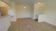 This single-floor space presents an open-concept layout that combines the living and dining areas into one cohesive, functional zone. The interior is finished with wall-to-wall neutral-toned carpeting that extends throughout, contributing to a warm and understated ambiance. Crown molding accents the upper perimeter of the space, adding a subtle architectural detail that enhances the overall finish quality.

A pendant light fixture is centrally positioned over what serves as the dining area, providing focused overhead lighting and serving as a modest focal point. Recessed lighting is also present, supplementing illumination throughout the space. The walls are painted in a clean, off-white tone that reflects light and maintains a bright, open feel.

A dedicated entryway is situated to the right, featuring a classic six-panel door with decorative glass inserts that allow natural light to filter into the foyer area. A half-wall partition subtly defines the entry zone from the main living space. A utility panel or HVAC vent is visible on the left wall, indicating practical climate control integration. Overall, the space offers a clean, move-in ready environment with straightforward, functional design.