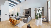 Located on Outlet Blvd in Fort Worth, TX, this single-floor community clubhouse delivers a sophisticated, modern aesthetic defined by soaring ceilings, exposed dark beam accents, and wide-plank light hardwood flooring throughout. The expansive main living area serves as the heart of the space, anchored by a striking floor-to-ceiling dark tile fireplace surround with a sleek linear hearth and wall-mounted TV. Floating wooden shelves with curated décor flank the fireplace, while a built-in marble-backed nook adds refined detail. The seating arrangement accommodates generous gatherings, complemented by a warm mix of neutral upholstery and cognac leather accents. The kitchen area is equally impressive, featuring dark gray cabinetry with gold hardware, a crisp marble tile backsplash, light stone countertops, glass-front upper cabinets, and a built-in microwave. A substantial dark wood island with an additional prep table anchors the kitchen, illuminated by large globe pendant lights in brushed nickel and brass. A separate lounge area near the entrance is bathed in natural light through floor-to-ceiling windows and glass doors that open to an outdoor balcony, framed by elegant patterned drapery. The overall ambiance is polished, welcoming, and well-suited for community gatherings.