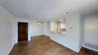 This single-floor space offers an open and functional layout designed for comfortable everyday living. The main living area features rich, dark hardwood-style flooring that extends throughout, providing warmth and visual continuity across the space. Neutral light gray walls create a clean, versatile backdrop, while a ceiling fan centered overhead ensures year-round comfort and air circulation.

A dark wood entry door with decorative paneling serves as a welcoming focal point at the front of the space. The living area flows naturally toward the kitchen, which is accessible via a pass-through opening in the wall — a design choice that enhances the open feel while maintaining a sense of defined zones. The kitchen is outfitted with white upper cabinetry, recessed lighting, and a granite-topped counter that doubles as a breakfast bar with a built-in sink. The lighter tile flooring in the kitchen contrasts subtly with the living area, further distinguishing the two spaces. Overall, the layout is practical and well-connected, making it well-suited for both daily routines and casual entertaining.