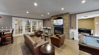 The Club Room at 500 Bedford St in Stamford, Connecticut is a thoughtfully designed amenity space that offers a variety of recreational and social areas across a single floor. The main lounge area serves as the heart of the space, featuring rich leather seating arranged around a striking stone-accented fireplace topped with a large flat-screen television. Light wood-style flooring, recessed lighting, and classic wainscoting throughout lend the space a refined yet comfortable character. Elegant French doors with glass panels connect the lounge to additional areas, allowing natural light to flow freely and creating a sense of openness. A dedicated game room provides ample entertainment options, housing a full-size pool table, a shuffleboard table, and a foosball table — making it an ideal gathering spot for friendly competition. A dining area with high-top seating offers a comfortable spot for casual meals or group gatherings. The fully equipped kitchen features rich cherry wood cabinetry, granite countertops, stainless steel appliances including a gas range and refrigerator, and a double sink beneath a window with natural light. A restroom is conveniently accessible off the kitchen area. Together, these well-appointed spaces make the Club Room a versatile and inviting amenity for residents and guests alike.