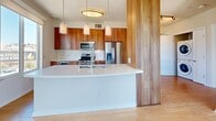 Located in the heart of San Diego, CA, this well-appointed two-bedroom, two-bathroom residence offers a thoughtfully designed single-floor layout. Light hardwood flooring flows throughout the main living areas, complemented by neutral tones and abundant natural light streaming through large windows and glass doors that frame city views.

The open-concept kitchen is a standout feature, showcasing rich warm-toned wood cabinetry, a sweeping white countertop island with pendant lighting, and a full suite of stainless steel appliances including a gas range and French door refrigerator. A structural wood column adds a natural architectural accent between the kitchen and living area.

The living room benefits from floor-to-ceiling windows and a ceiling fan, inviting ample daylight and urban views. Both bedrooms are generously sized, with one featuring hardwood floors and a city-facing window, while the other is finished with plush carpet and access to natural light.

The two bathrooms are distinctly appointed: one features large-format tile flooring, a glass-enclosed walk-in shower with pebble stone flooring, and warm wood cabinetry; the other offers a clean tiled tub-shower combination with matching cabinetry. A hallway with dedicated closets and in-unit laundry further enhances the practicality of this modern San Diego residence.