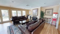 This single-story space centers around a spacious and well-appointed living area that combines comfort with functionality. The room features warm beige walls accented by crown molding, a textured ceiling, and hardwood flooring that adds a classic, inviting feel throughout.

The seating arrangement consists of two large dark brown leather sofas positioned around a dark wood coffee table, complemented by a decorative area rug with blue and floral patterning. A white wainscoted fireplace serves as a focal point along one wall, topped with a framed landscape painting that adds an artistic touch to the space.

Natural light flows generously into the room through a set of large windows along the front wall, offering views of mature trees and an outdoor area. A tall decorative faux plant adds a touch of greenery near the fireplace surround.

Toward the rear of the living area, a dedicated workspace includes a desk and computer setup, neatly integrated without disrupting the overall living space aesthetic. Additional framed artwork adorns the walls, contributing to the room's polished, residential character. The overall ambiance is warm, functional, and well-organized.