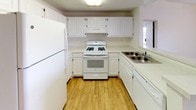 This single-floor space offers a clean, functional layout designed with practicality and comfort in mind. The kitchen serves as a standout feature, showcasing classic white raised-panel cabinetry that extends from floor to ceiling, providing generous storage throughout. White appliances, including a gas range with oven, a full-size refrigerator, and a dishwasher, are seamlessly integrated into the design, creating a cohesive and tidy aesthetic. The double-basin stainless steel sink is positioned beneath a window, allowing natural light to brighten the workspace. Light-toned countertops complement the white cabinetry, while a range hood overhead adds both function and a finished look to the cooking area. Warm, honey-toned hardwood-style flooring runs throughout the kitchen, adding a welcoming contrast to the crisp white surroundings and lending the space a sense of warmth. The U-shaped kitchen layout is thoughtfully designed to maximize efficiency and ease of movement, making it well-suited for everyday cooking and meal preparation. Overall, the space presents a bright, well-maintained environment with a timeless, neutral palette that offers a versatile and inviting atmosphere.
