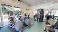 This fitness center offers a comprehensive workout facility spread across a single level, seamlessly blending an indoor gym with a thoughtfully designed outdoor exercise and recreation area. The interior gym is bright and airy, featuring large windows that flood the space with natural light and provide views of the surrounding greenery. Recessed lighting complements the natural light, illuminating a durable teal rubberized floor. The space is equipped with a full lineup of Precor cardio machines, including ellipticals and a stationary bike, alongside a multi-station strength training cable machine. Floor-to-ceiling mirrors along one wall visually expand the space and allow users to monitor their form during workouts. Transitioning outdoors, a covered patio area extends the fitness experience with Peloton spin bikes positioned beneath a sleek angled roof with warm cedar wood accents. Adjacent to the spin area, a sparkling swimming pool provides an additional avenue for exercise and relaxation. A furnished lounge patio with wicker and upholstered seating, string lights, and lush hedge privacy screens creates an inviting outdoor retreat. The overall ambiance balances functionality with comfort, making this a versatile and well-appointed fitness and recreation destination.