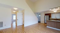 This two-story space makes a strong first impression with its dramatic vaulted ceiling in the main living area, creating an airy and expansive atmosphere. Light-toned hardwood flooring runs throughout the main level, lending warmth and continuity to the open layout. The generous ceiling height draws the eye upward, emphasizing the sense of volume and space.

The main living area flows naturally toward the entryway, which features a classic six-panel white door and an arched transom window above the front entrance, allowing natural light to filter in. The clean white walls throughout provide a bright, neutral backdrop adaptable to various interior styles.

Visible from the living area, the kitchen is partially open to the main space and includes a refrigerator, offering a functional layout. A staircase is integrated into the living area, with its diagonal line adding an architectural element to the room.

Multiple doorways and passage points suggest a well-connected floor plan across both levels. The overall ambiance is clean, bright, and move-in ready, with simple finishes that provide a versatile foundation for personalization.