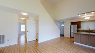 This two-story space makes a strong first impression with its dramatic vaulted ceiling in the main living area, creating an airy and expansive atmosphere. Light-toned hardwood flooring runs throughout the main level, lending warmth and continuity to the open layout. The generous ceiling height draws the eye upward, emphasizing the sense of volume and space.

The main living area flows naturally toward the entryway, which features a classic six-panel white door and an arched transom window above the front entrance, allowing natural light to filter in. The clean white walls throughout provide a bright, neutral backdrop adaptable to various interior styles.

Visible from the living area, the kitchen is partially open to the main space and includes a refrigerator, offering a functional layout. A staircase is integrated into the living area, with its diagonal line adding an architectural element to the room.

Multiple doorways and passage points suggest a well-connected floor plan across both levels. The overall ambiance is clean, bright, and move-in ready, with simple finishes that provide a versatile foundation for personalization.