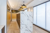 Located at 4401 Wornall Rd in Kansas City, Missouri, this mail and package room is a thoughtfully designed amenity space that balances functionality with upscale aesthetics. The room is anchored by a striking marble-clad island housing a full bank of individual mailboxes, finished in matte black and illuminated by a row of bold gold and black pendant lights overhead. The herringbone-patterned light wood flooring adds warmth and texture throughout the space, complementing the clean white and marble tones. A built-in marble-topped sorting counter with integrated drop slots provides a convenient surface for handling deliveries, and is accented by a curated gallery of contemporary framed artwork displayed on the adjacent wall. Along one side of the room, a full wall of modern package lockers in a neutral finish offers secure storage for larger deliveries. Floor-to-ceiling windows fitted with sheer roller shades allow diffused natural light to fill the space while maintaining privacy. The room connects seamlessly to a broader lobby area, which features soaring ceilings, rich dark wood architectural elements, large-format tile flooring, and a comfortable lounge with curated furnishings — reflecting the polished, contemporary character of the overall building.