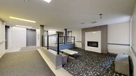 Located at 2700 Saratoga Pl in Ottawa, Ontario, this Homestead-branded residential lobby offers a well-appointed and functional entrance experience. The space is designed with a split-level layout, where a raised entry zone transitions down to a lower lounge area via steps bordered by sleek black metal railings with glass panels. The flooring is a striking combination of patterned dark terrazzo-style tile in the lounge area and light tile near the entry, creating a clear visual distinction between zones. A standout feature is the wall-mounted linear fireplace, framed in white with warm wood-panel accents, providing a welcoming focal point. Seating is provided through minimalist white bench-style tables and a dark wood bench positioned near a window with an exterior view of the surrounding landscape. The lobby is served by multiple elevators and features a secure glass-panelled entry vestibule with an intercom panel. Overhead, recessed linear LED lighting and ceiling-mounted spot fixtures ensure the space is well-lit throughout. White brick accent walls and neutral painted surfaces contribute to a clean, contemporary aesthetic. The overall ambiance is modern, organized, and inviting — designed to make a strong first impression for residents and visitors alike.