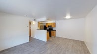 This single-floor space offers a practical and well-organized open-concept layout that connects the kitchen and living area in a cohesive, functional design. The kitchen is defined by a half-wall peninsula that serves as a natural divider, providing counter seating and a clear sightline into the main living space. Warm honey-toned wood cabinetry lines the kitchen walls, complemented by dark countertops and a full suite of stainless steel appliances, including a refrigerator, microwave, and range. The light gray laminate wood-style flooring runs throughout the space, lending a clean and contemporary feel underfoot. White walls and a white ceiling create a bright, neutral backdrop that maximizes the sense of openness. Recessed and flush-mount ceiling lighting provides consistent illumination throughout. A hallway visible beyond the kitchen peninsula suggests additional rooms, hinting at a well-connected floor plan. The overall ambiance is clean, modern, and move-in ready, with a straightforward design that prioritizes functionality and ease of living.