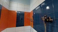 This single-floor utility wash station is thoughtfully designed to accommodate two specific functions: a dedicated dog wash area and a bike wash area. The space is immediately identifiable by its bold, eye-catching tilework, featuring a striking color palette of navy blue, orange, and white tiles arranged in horizontal bands across the walls. Each zone is clearly labeled with tiled text and iconography — a bicycle graphic marks the bike wash section, while paw print motifs distinguish the dog wash area, making the layout intuitive and easy to navigate. Multiple chrome faucet fixtures and handheld spray attachments are installed at practical heights throughout the space, ensuring both areas are fully equipped for thorough rinsing and cleaning tasks. The glossy ceramic tiles not only contribute to the space's vibrant aesthetic but also serve a highly functional purpose, offering durable, water-resistant, and easy-to-clean surfaces. The light-colored ceiling provides a clean contrast to the vivid walls below, keeping the space feeling bright and open. Overall, this wash station combines practicality with a playful, well-considered design, making it a standout amenity well-suited to pet owners and cycling enthusiasts alike.