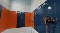 This single-floor utility wash station is thoughtfully designed to accommodate two specific functions: a dedicated dog wash area and a bike wash area. The space is immediately identifiable by its bold, eye-catching tilework, featuring a striking color palette of navy blue, orange, and white tiles arranged in horizontal bands across the walls. Each zone is clearly labeled with tiled text and iconography — a bicycle graphic marks the bike wash section, while paw print motifs distinguish the dog wash area, making the layout intuitive and easy to navigate. Multiple chrome faucet fixtures and handheld spray attachments are installed at practical heights throughout the space, ensuring both areas are fully equipped for thorough rinsing and cleaning tasks. The glossy ceramic tiles not only contribute to the space's vibrant aesthetic but also serve a highly functional purpose, offering durable, water-resistant, and easy-to-clean surfaces. The light-colored ceiling provides a clean contrast to the vivid walls below, keeping the space feeling bright and open. Overall, this wash station combines practicality with a playful, well-considered design, making it a standout amenity well-suited to pet owners and cycling enthusiasts alike.