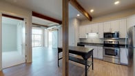 This single-floor unit showcases an appealing blend of industrial character and contemporary design. The open-concept living and kitchen area is immediately distinguished by exposed wooden beams and visible ductwork overhead, lending a loft-like aesthetic. Light wood-tone flooring runs throughout the main living space, complementing the neutral palette and enhancing the sense of openness. A sliding barn door adds a rustic architectural detail, while large windows flood the living area with natural light and provide outdoor views.

The kitchen is well-appointed with white shaker-style cabinetry, dark countertops, and a full suite of stainless steel appliances, including a refrigerator, range, and over-the-range microwave. A freestanding island with a dark work surface provides additional prep and dining space. The layout flows efficiently between the kitchen and living area, making it well-suited for both everyday living and entertaining.

The bedroom is a calm, carpeted retreat featuring a generously sized window that brings in natural light and exterior views. A dedicated walk-in closet with wire shelving offers practical storage. The bathroom is clean and functional, finished with wood-look flooring, a white vanity, and a full tub-shower combination.