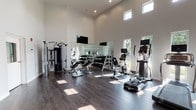 The fitness center at Halsey Flats is a well-appointed, single-level facility designed to support a comprehensive workout experience. The space is immediately distinguished by its dramatic double-height ceilings, which lend an open, airy atmosphere and contribute to an overall sense of spaciousness. Crisp white walls and dark hardwood-style flooring create a clean, contemporary aesthetic throughout.

Natural light enters through a series of clerestory windows positioned high along the walls, as well as larger windows at ground level that offer views of the surrounding greenery. This combination of natural and recessed overhead lighting ensures the space remains bright and inviting throughout the day.

The main workout area is divided into functional zones. A dedicated cardio section features multiple treadmills with integrated display screens, an elliptical trainer, and a stationary bike. The strength training area includes a multi-function cable machine, an adjustable weight bench, and a full dumbbell rack spanning a wide range of weights. Large wall-mounted mirrors run the length of one wall, enhancing the sense of depth and allowing users to monitor their form.

A mounted flat-screen television is positioned prominently for viewing during workouts. A separate entry area includes a storage credenza and water station, providing a functional transition point into the main floor.
