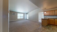 This single-floor residence offers a practical and comfortable layout with clearly defined living spaces. The open living area is generously proportioned, featuring vaulted ceilings that enhance the sense of space, neutral-toned carpet underfoot, and multiple windows that allow ample natural light to fill the room. A wall-mounted air conditioning unit and baseboard heating provide year-round climate control. The living area flows naturally toward the kitchen, which is separated by a raised breakfast bar. The kitchen is well-appointed with warm wood cabinetry, dark granite-style countertops, and a full suite of white appliances including a refrigerator, electric range, microwave, and dishwasher. Stone-look tile flooring extends through the kitchen and entry areas, adding durability and a cohesive aesthetic. The bedroom is a quiet, carpeted retreat with a large window and baseboard heating. The bathroom features a warm wood vanity with a stone-top countertop, a large mirror with overhead lighting, tile flooring, and a separate tub and shower area. The entryway, finished in matching tile, provides a welcoming transition into the home. Overall, the space is clean, functional, and well-maintained with a consistent neutral color palette throughout.