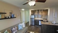 This single-floor residence offers a thoughtfully designed open-concept layout that seamlessly connects the living area and kitchen, creating a cohesive and functional living space. The kitchen is a standout feature, showcasing rich dark cabinetry, stainless steel appliances including a full-size refrigerator, and a decorative mosaic tile backsplash that adds texture and visual interest. A pendant light and track lighting overhead provide both task and ambient illumination, while light stone countertops contrast beautifully against the darker tones of the cabinetry. The living area flows naturally from the kitchen, offering a comfortable and intimate setting anchored by a ceiling fan with integrated lighting. Dark hardwood-style flooring runs throughout the space, lending warmth and continuity from room to room. A hallway visible from the main living area leads to additional rooms, suggesting a well-organized and practical floor plan. The overall ambiance is modern and inviting, with a harmonious blend of dark and light tones, contemporary fixtures, and quality finishes that come together to create a stylish and comfortable living environment.
