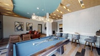 The game room is a well-appointed recreational space designed to balance active entertainment with relaxed social gathering. At its center sits a full-size pool table with a blue felt surface, finished in a sleek metallic frame and illuminated by a rectangular pendant chandelier that anchors the room visually. The ceiling is one of the space's most distinctive architectural features, combining a bold teal-painted section with a multi-tiered wood-paneled surround accented by warm recessed strip lighting, adding depth and a contemporary feel overhead. Light hardwood flooring runs throughout, contributing to a bright, open atmosphere. To one side, a comfortable lounge area features leather seating and bold abstract artwork on white walls, creating an inviting spot to unwind between games. On the opposite side, a built-in counter with seating provides a dedicated workspace or casual bar-style area. A small round table with modern chairs offers additional seating. The overall design blends modern aesthetics with warm materials, making this game room a versatile and visually engaging communal space.
