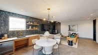 Located at 2805 S Cooper St in Arlington, Texas, this thoughtfully designed single-floor space blends contemporary aesthetics with functional communal living. The open layout seamlessly connects a dining area, lounge seating zone, and a built-in wet bar or kitchenette, creating a cohesive and inviting environment throughout.

The dining area centers on a sculptural round marble-topped table surrounded by cream upholstered chairs, illuminated by a modern brass multi-arm pendant light. Adjacent to it, a warm-toned wood kitchenette features gold hardware and marble countertops, complemented by a dramatic floor-to-ceiling wall clad in deep green glazed tile. Floating shelves above display curated décor, while a wall-mounted television is integrated seamlessly into the tile feature wall.

A double-sided fireplace with a bold matte black surround anchors the transition between the dining and lounge zones, adding warmth and visual drama. The lounge area is furnished with a cognac leather sofa, cream accent chairs, and layered rugs atop large-format stone-look tile flooring that flows throughout the space.

A dedicated banquette seating area features a continuous navy upholstered bench set against an eye-catching decorative wallcovering displaying an eclectic collage of framed artwork on a plaid background, lit by wall-mounted brass sconces. Recessed lighting throughout ensures a well-illuminated, polished ambiance.