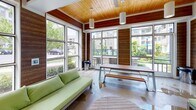 Located at 9550 Gannon Dr in Charlotte, NC, this single-floor lounge space offers a thoughtfully designed interior that blends natural materials with contemporary style. The room is defined by rich, horizontal wood-paneled walls that create a warm and textured aesthetic throughout, complemented by a striking natural wood-plank ceiling that adds depth and warmth overhead. Pendant cylinder light fixtures hang from the ceiling, providing soft, focused illumination that enhances the inviting atmosphere. Large, floor-to-ceiling multi-pane windows wrap around much of the space, flooding the interior with abundant natural light and offering open views of the surrounding landscaped grounds, trees, and neighboring buildings. The polished concrete floors add an urban, industrial contrast to the organic warmth of the wood elements. Seating includes vibrant green upholstered sofas and a wooden picnic-style table with bench seating, making the space functional for both relaxed lounging and casual gatherings. A wall-mounted flat-screen television serves as a focal point on one of the wood-paneled walls. The overall ambiance is bright, airy, and welcoming, successfully merging the comfort of an indoor retreat with a strong visual connection to the outdoors.