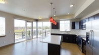 This contemporary single-floor space is defined by its open-concept layout and an abundance of natural light streaming through expansive floor-to-ceiling windows. The kitchen serves as the centerpiece of the space, showcasing a large central island with a clean white countertop that provides ample workspace and a natural gathering point. Sleek dark cabinetry lines the walls, offering generous storage while creating a striking contrast against the light-toned hardwood flooring and crisp white ceilings. Three bold red pendant lights hang above the island, adding a vibrant pop of color and serving as a dramatic design focal point. Recessed lighting throughout ensures the space remains well-illuminated at any time of day. A full-length glass sliding door opens directly onto a private balcony, seamlessly blending indoor and outdoor living while framing panoramic views of the surrounding cityscape and lush treetops. The open floor plan allows the kitchen and living areas to flow naturally into one another, creating a sense of spaciousness and versatility. Every detail, from the modern fixtures to the thoughtfully chosen finishes, reflects a cohesive and polished contemporary aesthetic throughout.