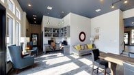 This single-floor space combines a functional work area with a comfortable lounge setting in an open, airy layout. The interior is defined by striking contrast between crisp white walls and a bold dark ceiling, lending the space a contemporary, polished aesthetic. Light wood-toned flooring runs throughout, brightening the room and complementing the neutral color palette.

A built-in workstation lines one wall, featuring an open shelving unit above two computer desks paired with ergonomic office chairs. The shelving provides both storage and display space, adding visual interest with decorative objects. Pendant lights in varying styles hang above the work area, providing focused task lighting while contributing to the room's eclectic design character.

Adjacent to the workstation, a lounge area offers a grey sofa dressed with yellow and patterned cushions, accompanied by additional seating. A circular red mirror and framed artwork adorn the walls, introducing bold accent colors. Recessed lighting supplements the natural light streaming in from a glass entrance door, ensuring the space remains bright and welcoming throughout the day.