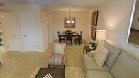 This single-story space features an open-plan layout that seamlessly connects the living and dining areas, creating a cohesive and functional environment. The living area is anchored by plush, light-toned carpeting and is furnished with a comfortable sofa accented with decorative pillows, a striped accent chair, and a dark wood coffee table. A shaggy area rug adds texture and warmth underfoot. Adjacent to the living area, the dining space is defined by a dark wood table paired with matching chairs, positioned atop a patterned area rug. A classic chandelier with warm-toned bulbs hangs above the dining table, serving as a focal point and providing ambient lighting. The walls are finished in a soft, neutral beige tone that enhances the overall warmth of the space. Decorative wall art, including a rustic wooden panel piece and framed geographic prints, adds visual interest. A side table with a table lamp provides additional lighting in the living zone, while a small plant introduces a natural element. The space benefits from a clean, tiled ceiling detail and consistent flooring throughout, contributing to a polished and well-composed interior.