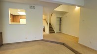 This two-story residence offers a straightforward and functional layout designed for comfortable everyday living. The main level greets visitors with a generously sized living area finished in neutral-toned carpet throughout, creating a warm and cohesive feel across the space. A notable architectural detail is the subtle split-level step within the living area, which adds dimension and visual interest to the open floor plan. Crisp white walls and a textured ceiling reflect light effectively, keeping the space feeling bright and airy. A central staircase, accented with wooden railings, serves as the natural focal point of the main level, providing a clean transition to the upper floor. Built-in shelving is visible near the staircase area, offering practical storage solutions. HVAC vents are integrated into the ceiling, ensuring climate control throughout the home. The open sightlines between areas of the main floor allow for a connected and spacious feel. The upper level, accessible via the carpeted staircase, extends the living space further. Overall, the home presents a clean, move-in ready canvas with a neutral palette and practical design elements suited to a variety of living arrangements.