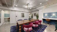 Located at 2400 Grayland Ave in Richmond, Virginia, this single-floor space offers a well-appointed and thoughtfully designed collaborative environment. The layout comprises two distinct areas that together create a functional yet visually dynamic setting.

The central meeting area is anchored by a substantial wooden farmhouse-style dining table with elegantly turned legs, surrounded by a mix of deep navy blue and rich magenta upholstered chairs. A bold navy and gold geometric-patterned area rug grounds the seating arrangement, while a striking brass Sputnik-style chandelier and exposed industrial track lighting illuminate the space from above. Polished concrete floors add an industrial edge throughout.

One wall features a sophisticated teal wainscoting panel installation with built-in shelving and sconce lighting, complemented by a built-in desk nook with individual task lighting — ideal for focused individual work. A frosted-glass double-door entry provides a sleek, professional entrance point with an adjacent private room accessible via a solid door.

Decorative touches including tall potted palms, white orchids, framed wall art, and curated accent furniture — including a tufted emerald green armchair and a sculptural round accent rug — contribute to the space's eclectic, inviting ambiance. Exposed ductwork along the ceiling reinforces the modern industrial character of the overall design.