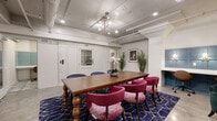 Located at 2400 Grayland Ave in Richmond, Virginia, this single-floor space offers a well-appointed and thoughtfully designed collaborative environment. The layout comprises two distinct areas that together create a functional yet visually dynamic setting.

The central meeting area is anchored by a substantial wooden farmhouse-style dining table with elegantly turned legs, surrounded by a mix of deep navy blue and rich magenta upholstered chairs. A bold navy and gold geometric-patterned area rug grounds the seating arrangement, while a striking brass Sputnik-style chandelier and exposed industrial track lighting illuminate the space from above. Polished concrete floors add an industrial edge throughout.

One wall features a sophisticated teal wainscoting panel installation with built-in shelving and sconce lighting, complemented by a built-in desk nook with individual task lighting — ideal for focused individual work. A frosted-glass double-door entry provides a sleek, professional entrance point with an adjacent private room accessible via a solid door.

Decorative touches including tall potted palms, white orchids, framed wall art, and curated accent furniture — including a tufted emerald green armchair and a sculptural round accent rug — contribute to the space's eclectic, inviting ambiance. Exposed ductwork along the ceiling reinforces the modern industrial character of the overall design.