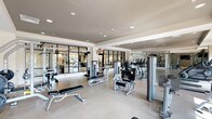 Located at 260 Leigh Farm Rd in Durham, NC, this professionally equipped fitness center offers a spacious, single-floor layout designed to accommodate a wide range of workout needs. The facility is bathed in natural light streaming through large floor-to-ceiling windows along the perimeter, complemented by recessed ceiling lighting that ensures a bright, energizing environment throughout.

The space is organized into distinct functional zones. A dedicated strength training area houses an extensive selection of selectorized resistance machines, free weight benches, and a full dumbbell rack, all set against floor-to-ceiling mirrors that enhance the sense of depth. Glass-panel partitions divide sections of the facility while maintaining an open, airy feel.

The cardio zone features a long row of treadmills, stationary bikes — including upright and recumbent models — ellipticals, and stair climbers, many equipped with integrated screens. Wall-mounted televisions are positioned throughout for entertainment during workouts. A functional training area includes a TRX suspension training rig, medicine balls, and balance equipment set on light hardwood-style flooring, ideal for dynamic and circuit-based training.

The overall aesthetic is modern and clean, with neutral tones, durable flooring, and a well-organized layout that supports both individual and group fitness routines. Outdoor views through the windows add a welcoming ambiance to the entire space.